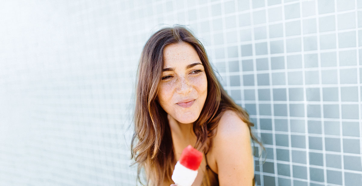 Eine junge Frau hält ein Eis am Stiel in der Hand und lächelt.
